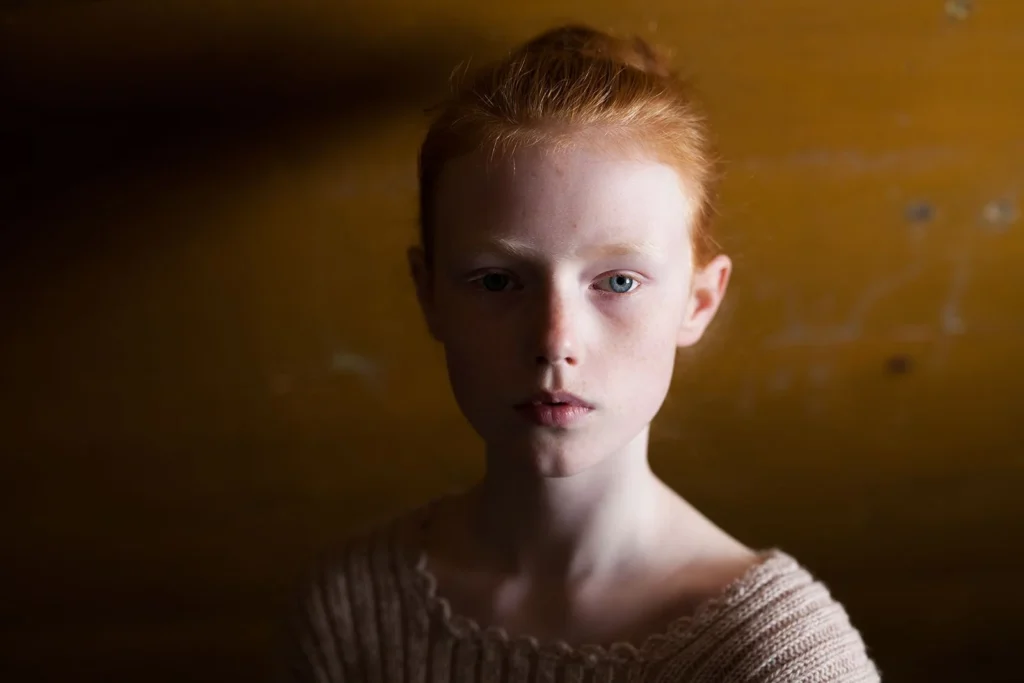 Retrato íntimo de joven pelirroja con moño, fondo ocre texturizado y luz dramática lateral.