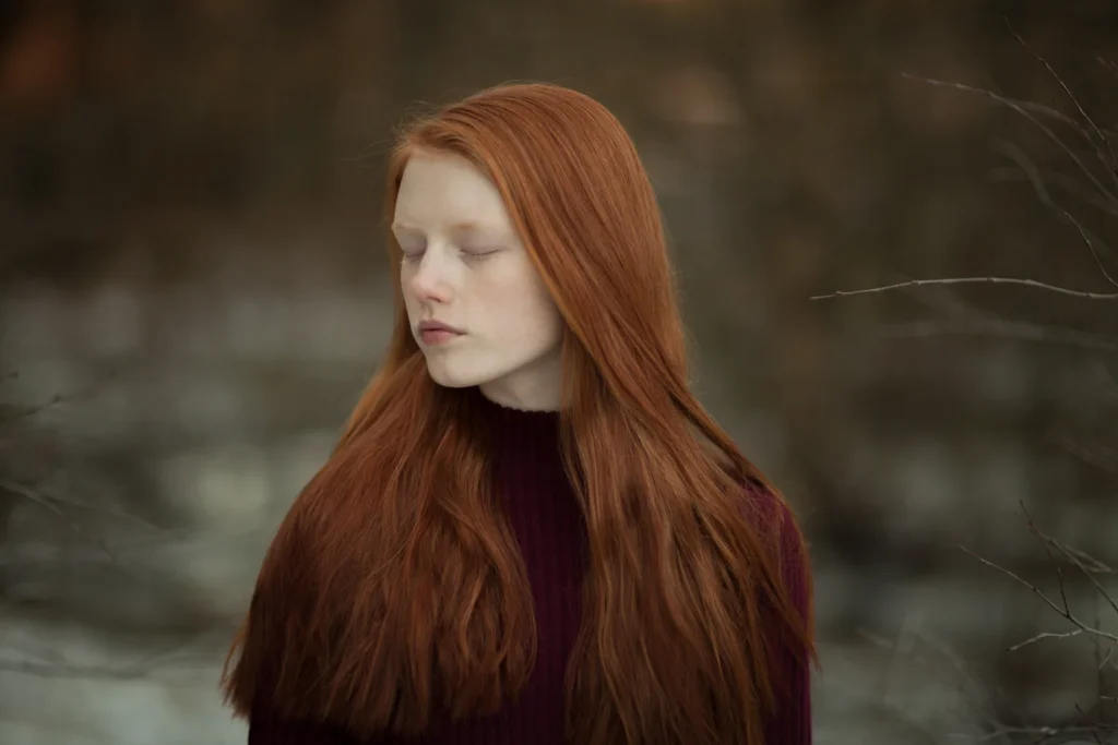 Joven pelirroja con ojos cerrados, suéter burdeos, fondo natural difuminado y luz suave.