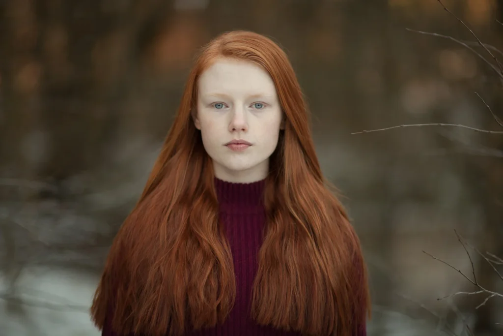 Joven pelirroja con suéter burdeos, mirada directa, fondo natural difuminado y luz suave.