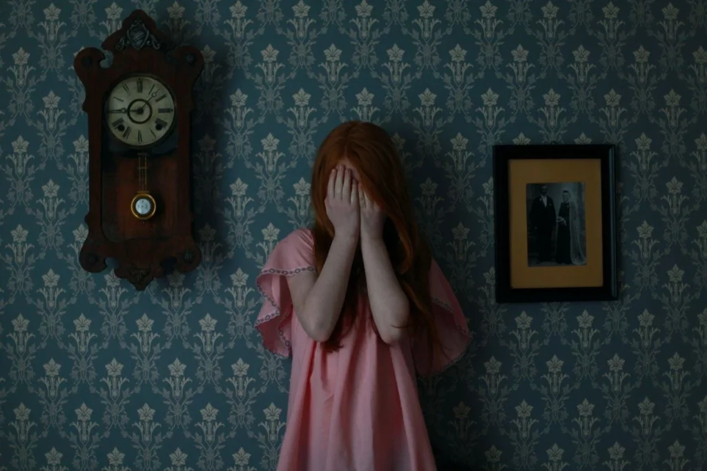 Joven pelirroja con vestido rosa cubriéndose el rostro, fondo con reloj antiguo y fotografía enmarcada.