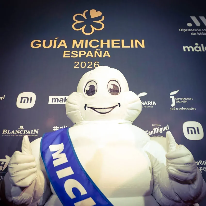 El muñeco Michelin sonriente con banda azul frente al cartel oficial de la Guía Michelin España 2026.