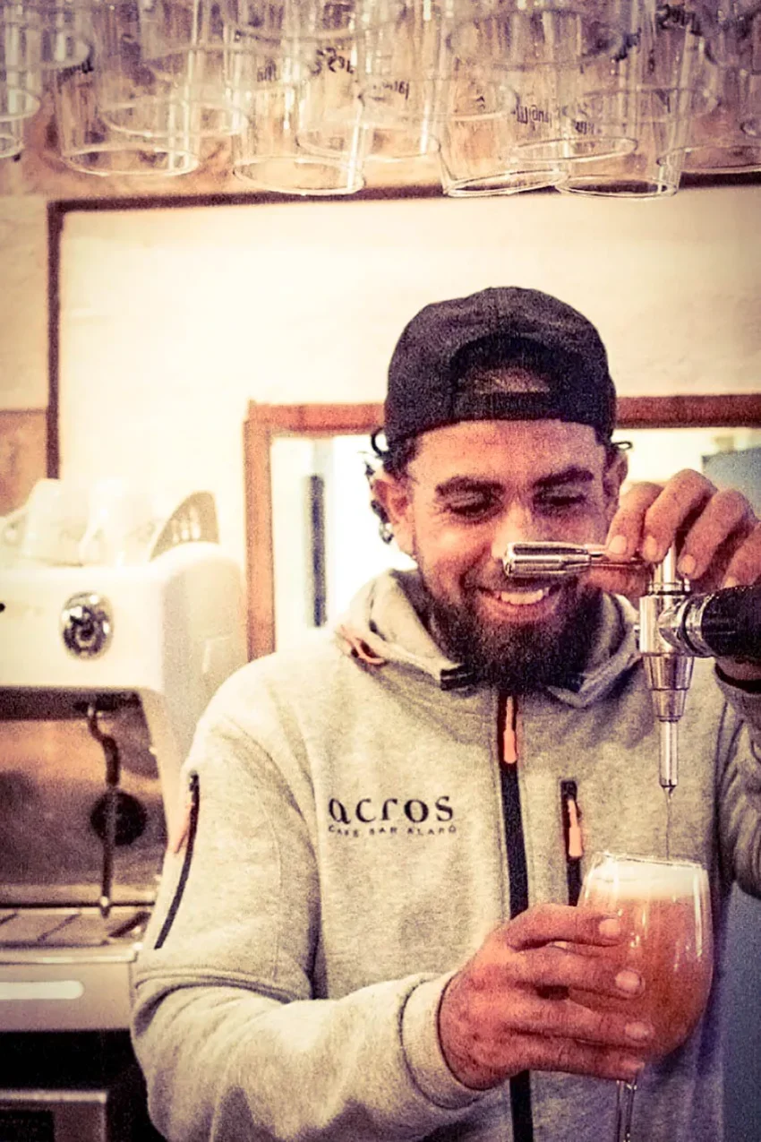 Joan Chochin sirviendo una bebida en el Acros Café del Rapé, con sudadera gris y gorra negra, sonriente y relajado.