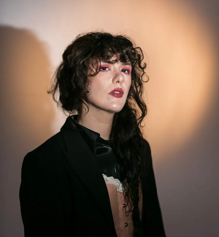 Retrato artístico de Raisa Maudit con maquillaje expresivo, chaqueta negra y fondo iluminado en tonos cálidos.