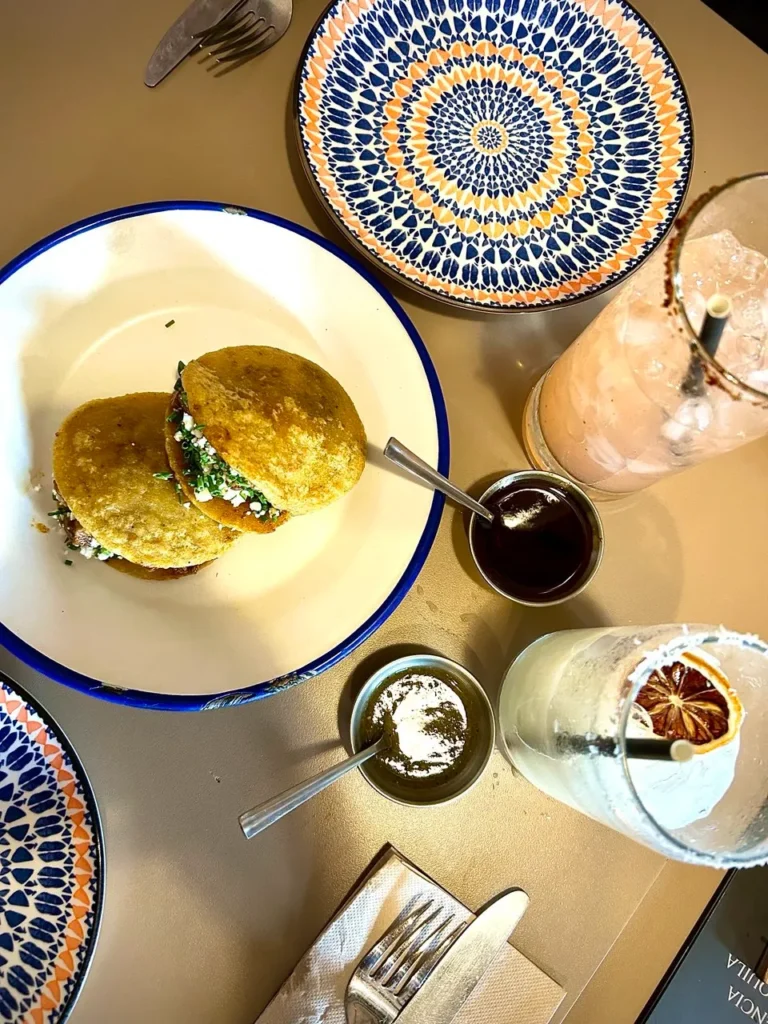 Dos gorditas doradas rellenas de queso, verduras y carne, servidas en un plato blanco con borde azul, acompañadas de salsas y bebidas coloridas sobre una mesa elegante.