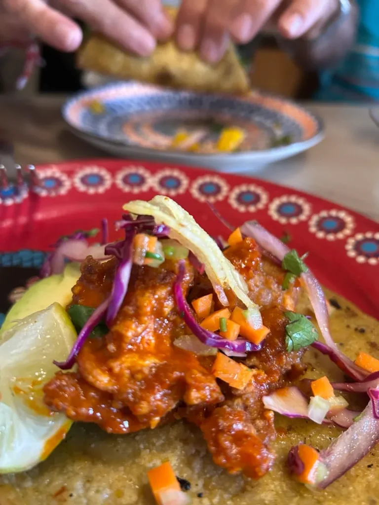 Taco servido en plato rojo decorativo, con carne sazonada, verduras frescas y una rodaja de lima, en un entorno de comida compartida.