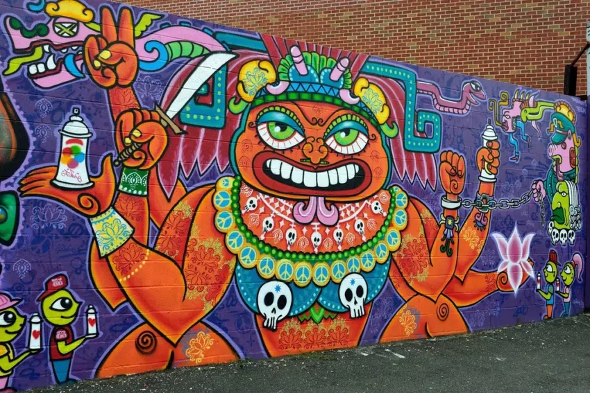 Mural de Lady Pink con figura antropomórfica multibrazo, símbolos culturales y personajes cartoon