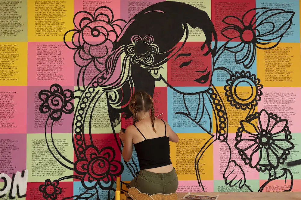 Artista pintando mural con figura femenina, flores y fondo de bloques de texto multicolor