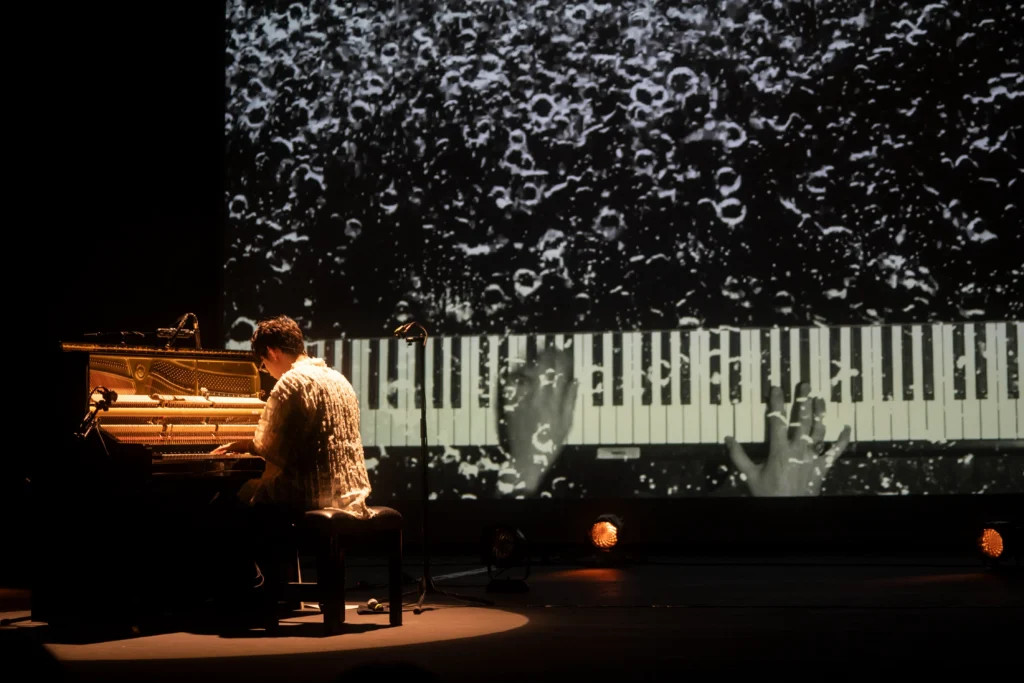 Pianista tocando un piano abierto en un escenario iluminado, con una proyección en blanco y negro mostrando teclas y efectos visuales durante el concierto.