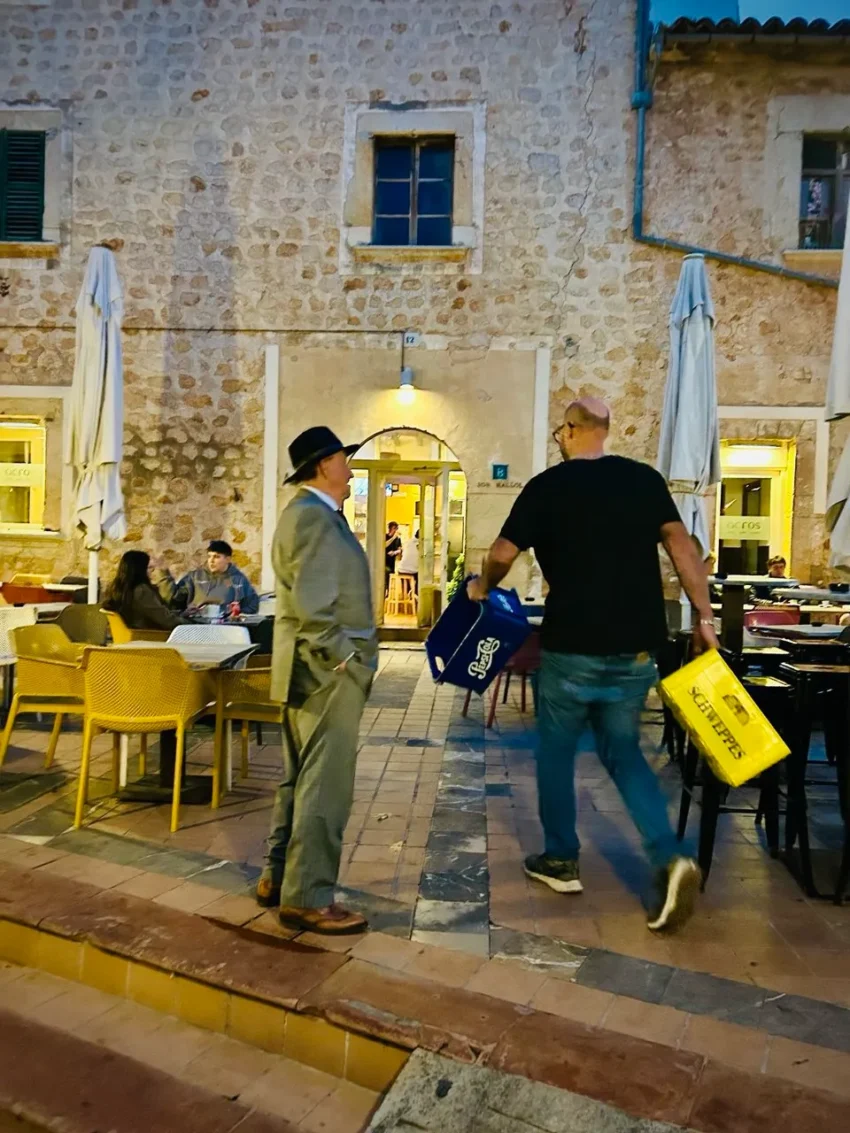 Escena en una terraza mediterránea al atardecer con dos hombres conversando mientras otro pasa cargando cajas de bebidas.
