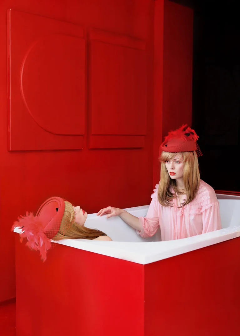 Dos mujeres con sombreros rojos en bañera blanca, en escena teatral sobre fondo rojo geométrico.