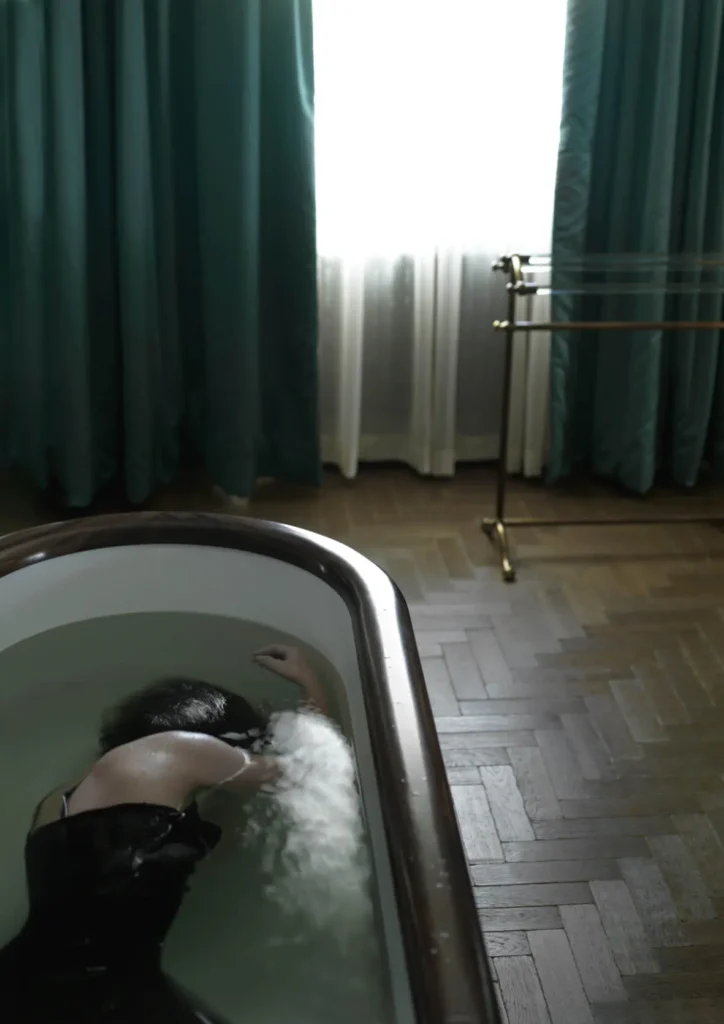 Mujer vestida de oscuro sumergida en una bañera, iluminada por luz natural, en escena introspectiva y teatral.