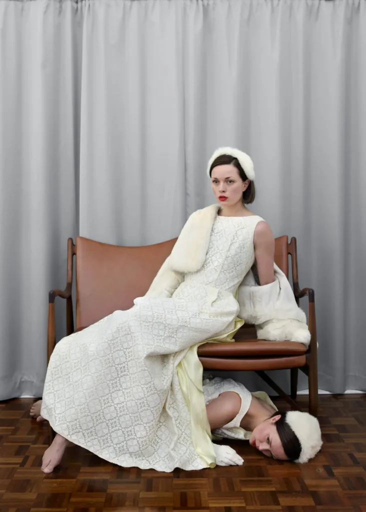 Dos mujeres vestidas de blanco con accesorios de piel, una sentada y otra oculta bajo el banco, en escena surrealista.