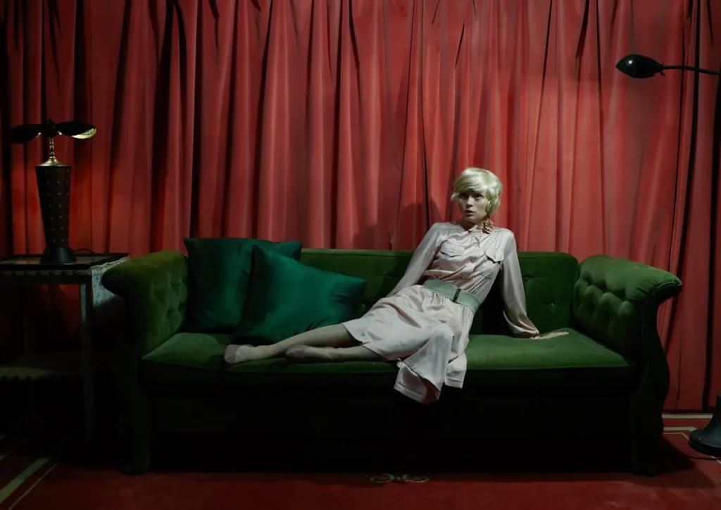 Mujer rubia recostada en sofá verde, con vestido rosa, frente a cortinas rojas en escena teatral.