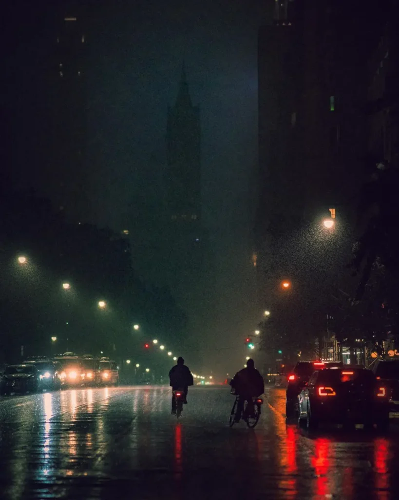 Fotografía de Nicolas Miller de dos ciclistas nocturnos en una calle mojada de Nueva York, con luces y sombras.