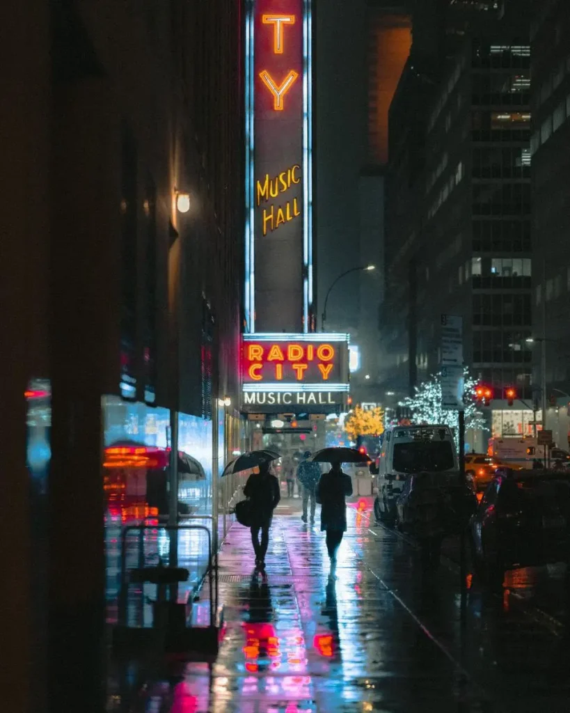 Fotografía de Nicolas Miller de Radio City Music Hall en Nueva York, con neón rojo, lluvia y figuras con paraguas.