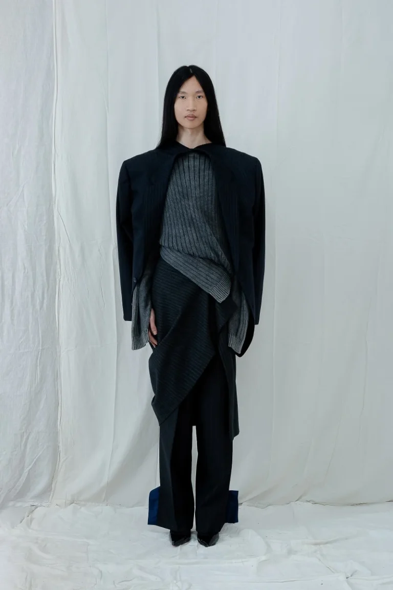 Modelo con look de Antonio Acuario: blazer estructurado, suéter gris y falda asimétrica sobre pantalón, frente a fondo claro.