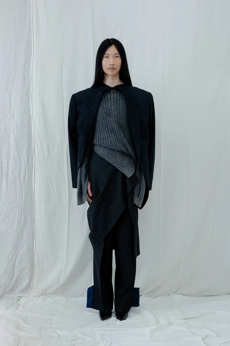 Modelo con look de Antonio Acuario: blazer estructurado, suéter gris y falda asimétrica sobre pantalón, frente a fondo claro.