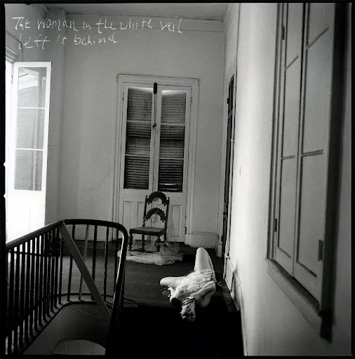 Figura femenina en traje blanco tendida frente a puertas con cortinas, escena surrealista en blanco y negro