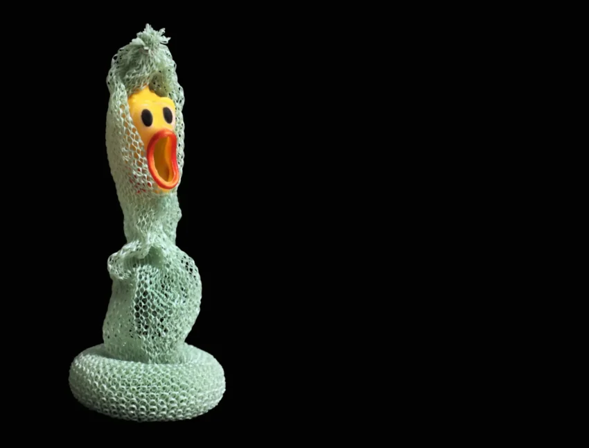 Juguete de pato envuelto en material verde texturizado sobre fondo negro, imagen surrealista y provocadora.