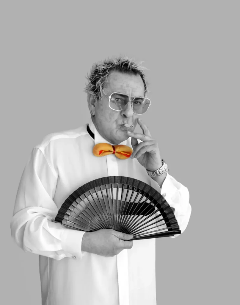 Carlos Penas con pajarita naranja, abanico negro y cigarro, en retrato monocromo con acento cromático.