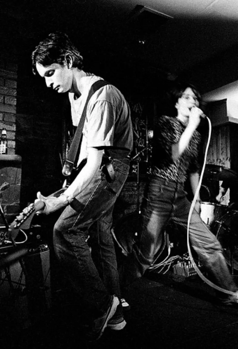 Life Without Buildings en directo: guitarrista concentrado y vocalista enérgica en un concierto íntimo, con batería al fondo y muro de ladrillo.