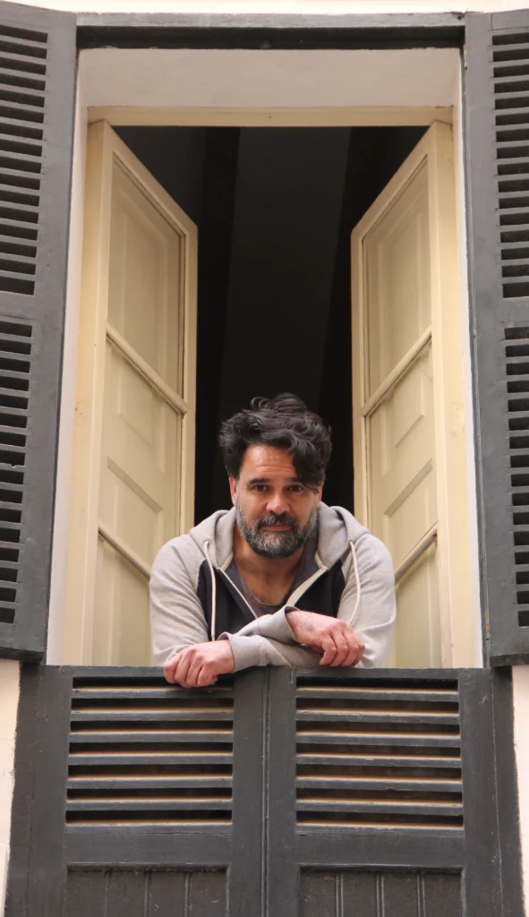 Llorenç Garrit con sudadera gris apoyado en la barandilla de una ventana abierta, en actitud contemplativa.