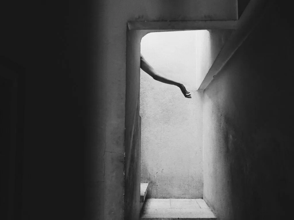 Brazo extendido que asoma desde un marco superior en una escalera oscura de hormigón, creando una escena minimalista y enigmática propia de la estética conceptual de Carolina Mateu.