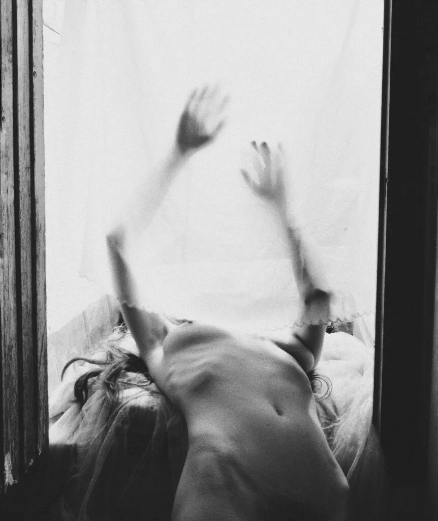 Escalera oscura de hormigón iluminada desde arriba, con un brazo extendido que asoma por una abertura superior, en una composición minimalista y enigmática de Carolina Mateu.