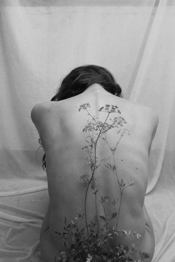 Espalda desnuda de una persona sentada y ligeramente inclinada hacia adelante, con una superposición artística que integra formas vegetales a lo largo de la columna en una escena en blanco y negro.