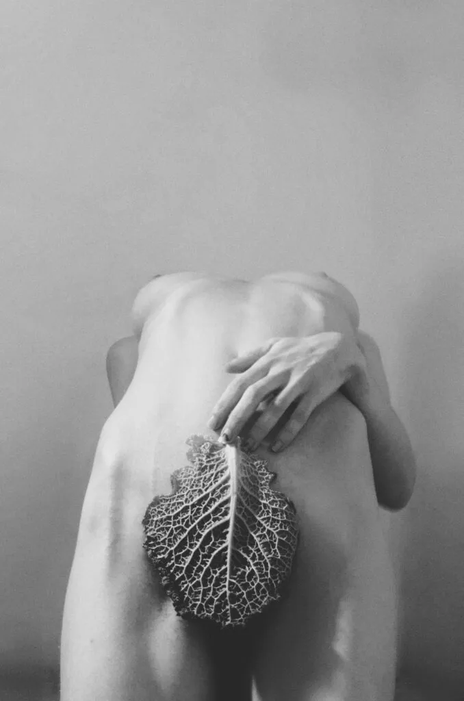 Espalda desnuda de una persona inclinada hacia adelante, con una intervención visual que integra motivos vegetales a lo largo de la columna en una escena en blanco y negro.