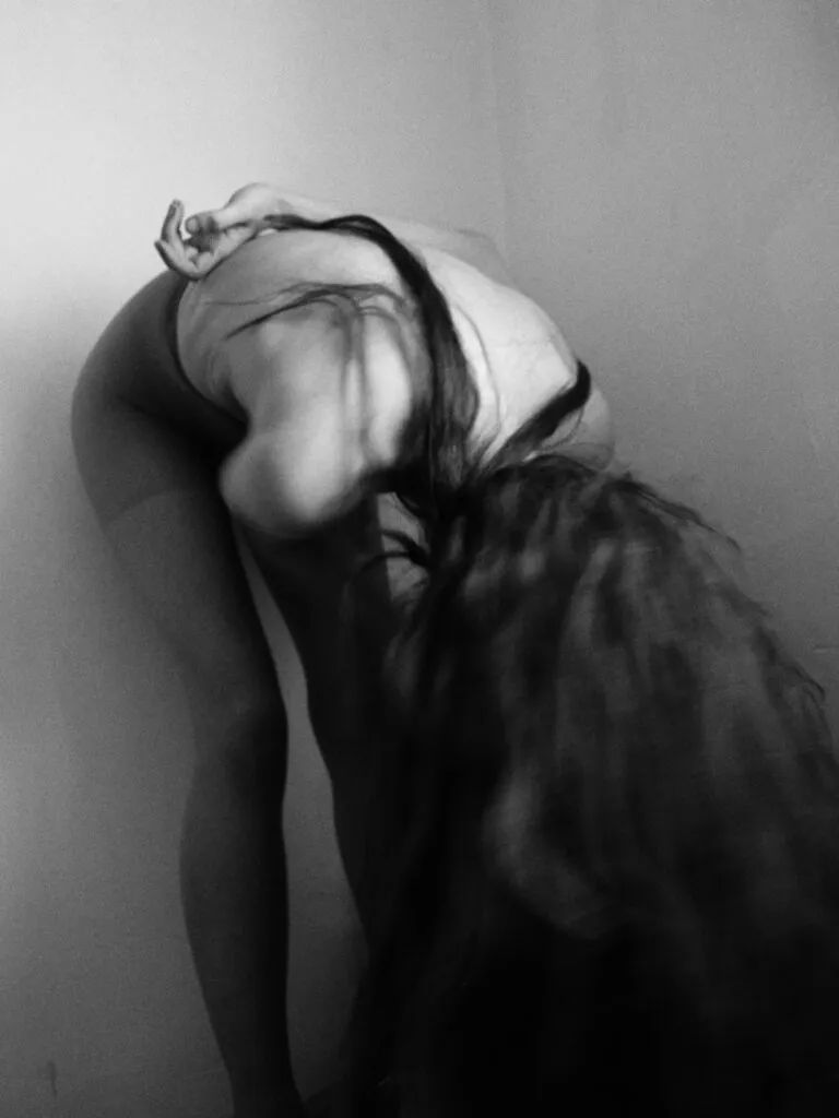 Persona en una pose contorsionada, inclinada hacia adelante con el cabello cayendo y los brazos tensos detrás del cuerpo, en una esquina iluminada suavemente en blanco y negro.