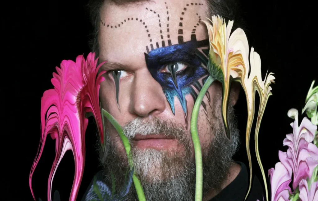 Primer plano artístico de John Grant con maquillaje expresivo y flores surrealistas alrededor.