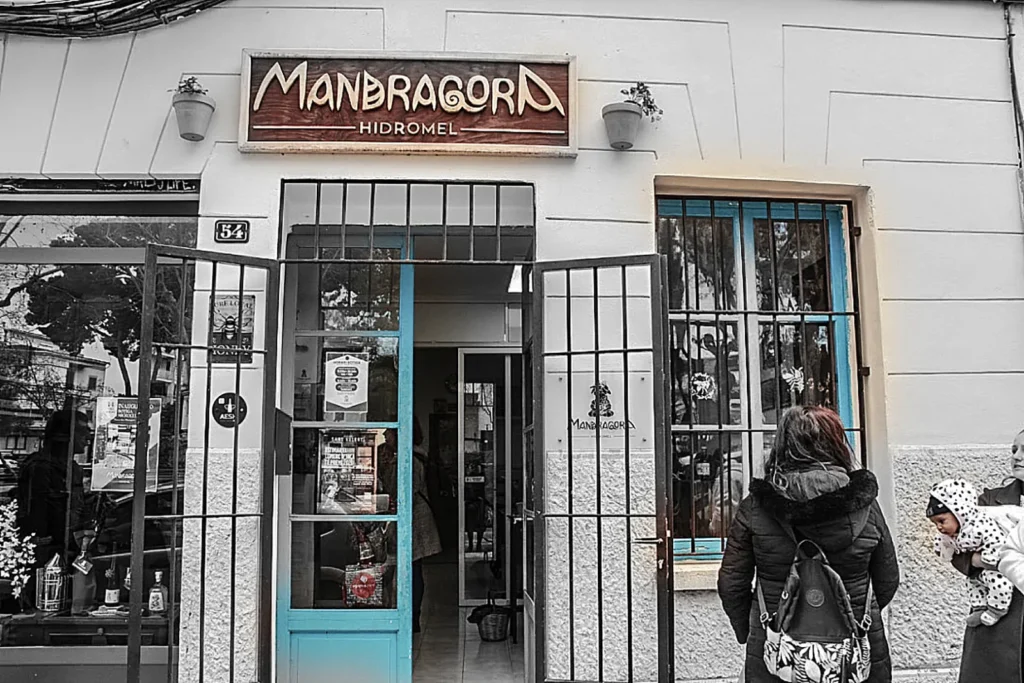 Fachada de Mandrágora Hidromel con rótulo de madera y entrada azul en una escena parcialmente desaturada.