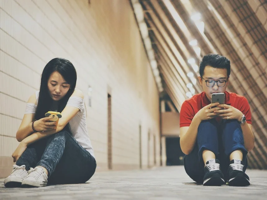 Dos personas sentadas en un pasillo moderno mirando sus teléfonos móviles, absortas en sus pantallas.