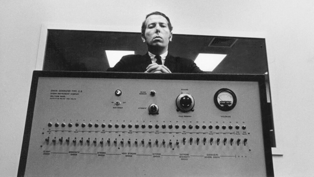 “Panel del ‘Shock Generator Type ZLB’ del experimento de Milgram, con un hombre de pie detrás del dispositivo y una serie de interruptores etiquetados con niveles crecientes de descarga eléctrica.”