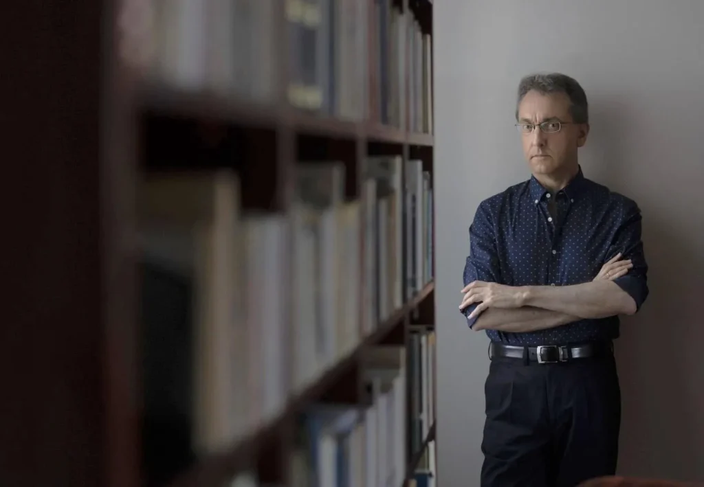 Jesús G. Maestro posando con los brazos cruzados junto a una estantería llena de libros.