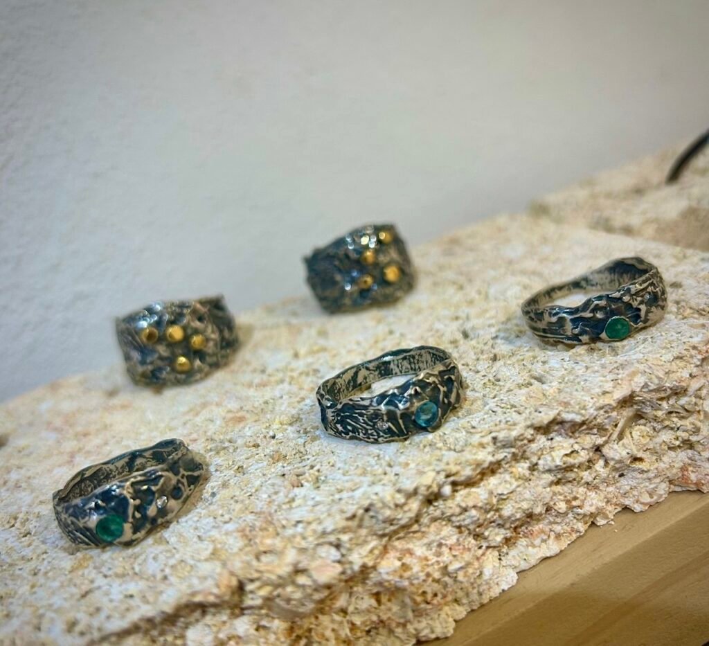 Cinco anillos artesanales de Rako Joies en metal oscuro con incrustaciones doradas y turquesa, dispuestos sobre una superficie beige.