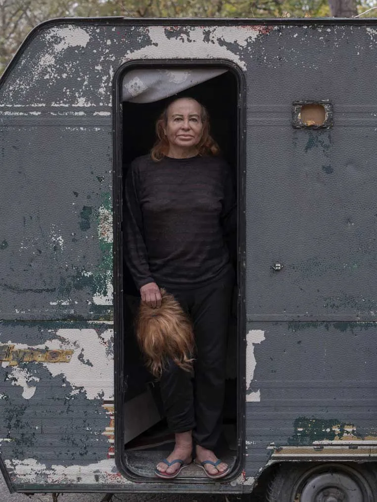 Una persona de mediana edad de pie en el marco de la puerta de una caravana antigua y desgastada, sosteniendo una peluca de color castaño.