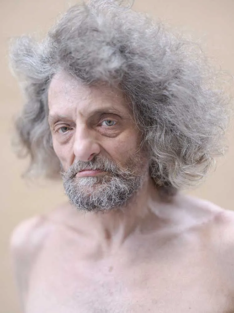 Primer plano de un hombre mayor con cabello largo y canoso, barba gris y torso descubierto, mirando fijamente a cámara sobre un fondo claro desenfocado.