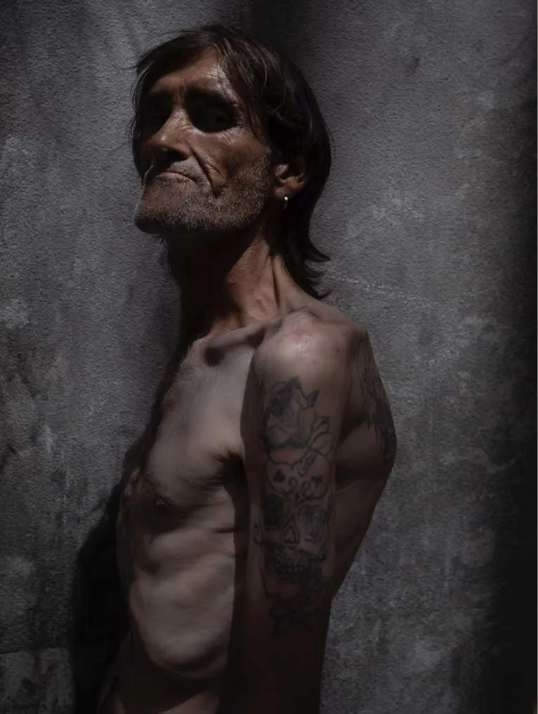 Retrato de perfil de un hombre delgado con el torso descubierto y tatuajes en el brazo, iluminado lateralmente con sombras profundas sobre un fondo de pared de cemento.