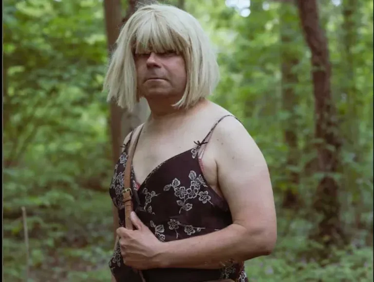 Retrato de plano medio de una persona con peluca rubia con flequillo y vestido de flores, posando en medio de un bosque verde frondoso.