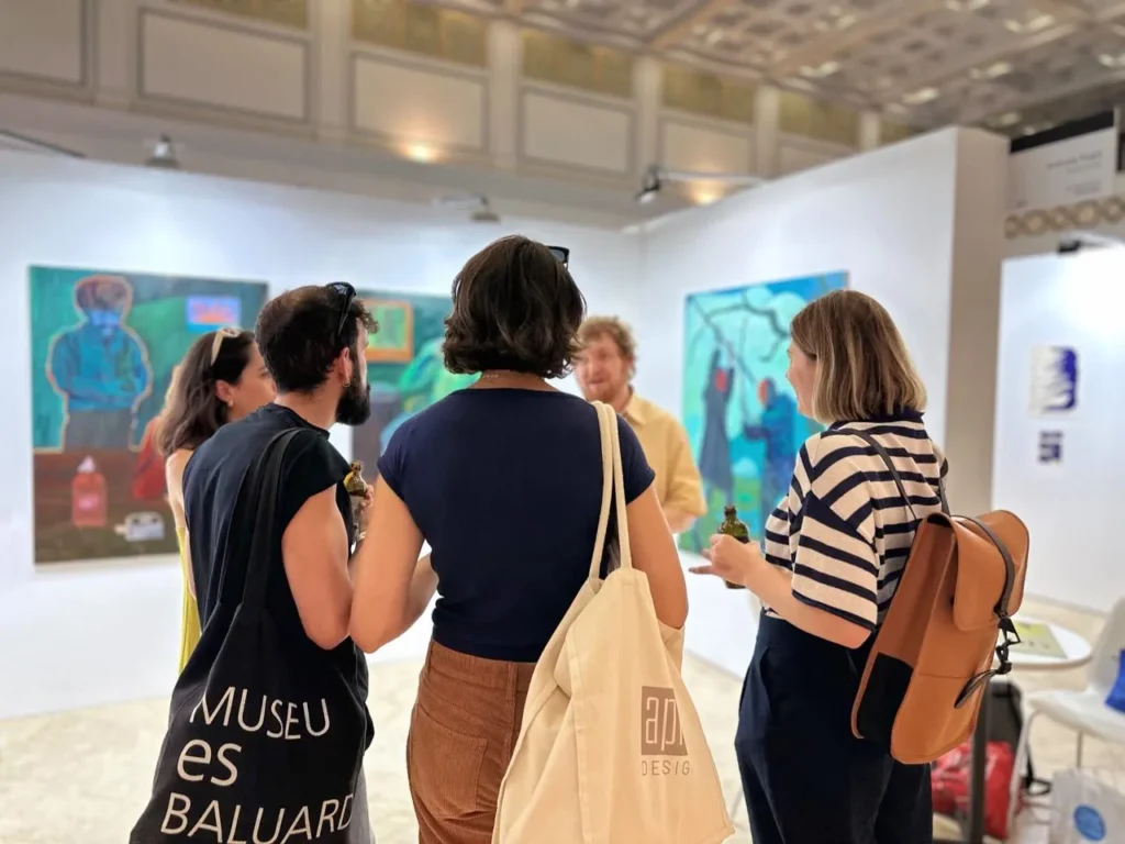 Grupo de cinco personas conversando en una galería de arte, rodeados de pinturas coloridas y un ambiente expositivo contemporáneo.