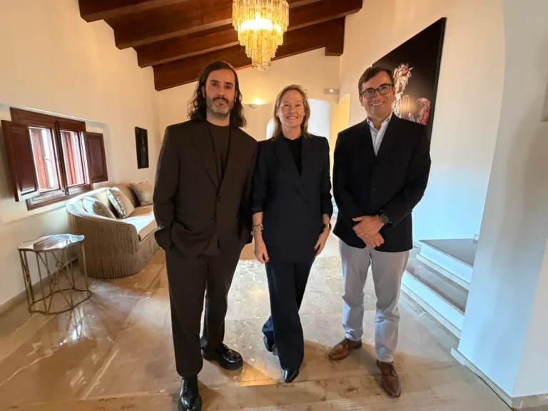 Tres personas posan en una sala elegante con vigas de madera, un sofá y una escalera al fondo.