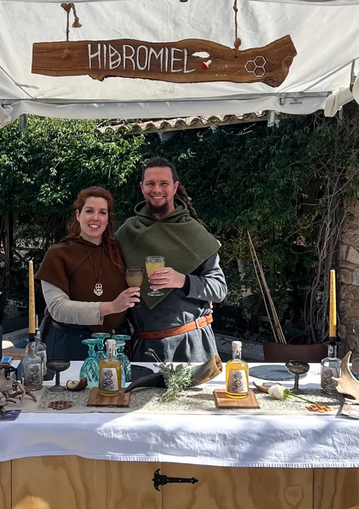Puesto de hidromiel Mandragora con dos personas vestidas de época atendiendo una mesa al aire libre.