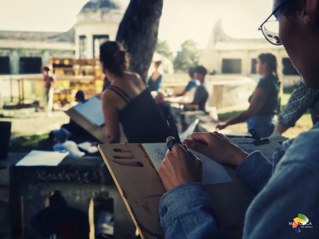 “Participantes de Quarantine Menorca realizando dibujo al aire libre en la isla de Lazareto, frente a arquitectura histórica.”