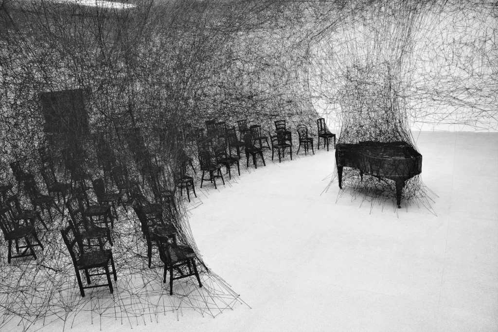 La firma distintiva de Chiharu Shiota son sus instalaciones con hilos de lana, principalmente en colores rojo intenso y negro. Estos elementos no son meros componentes decorativos, sino verdaderas metáforas visuales que conectan: