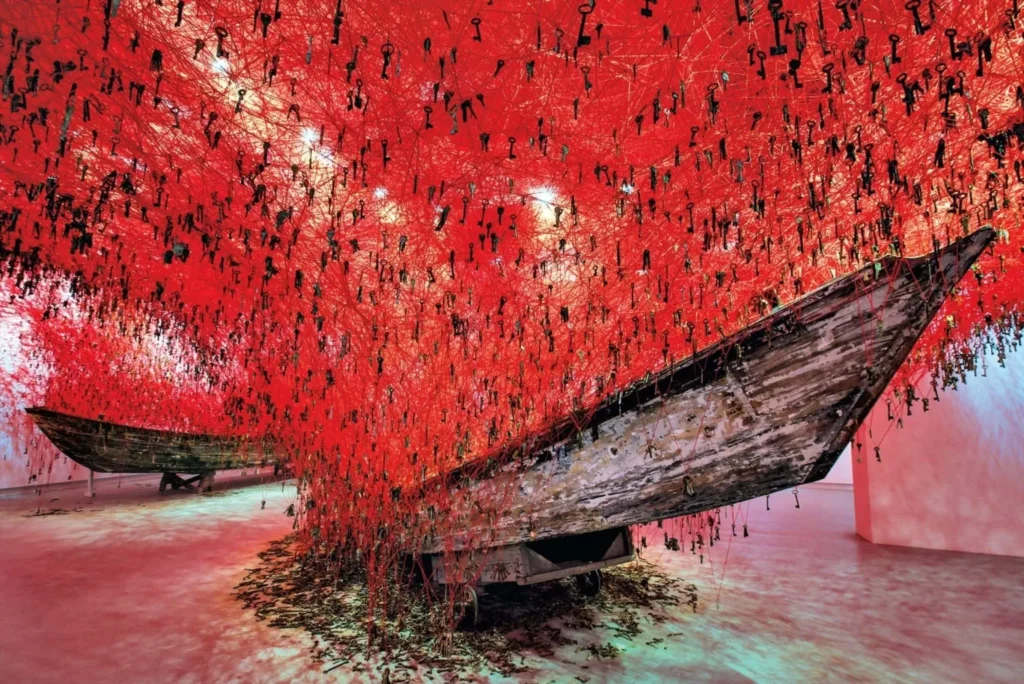 La firma distintiva de Chiharu Shiota son sus instalaciones con hilos de lana, principalmente en colores rojo intenso y negro. Estos elementos no son meros componentes decorativos, sino verdaderas metáforas visuales que conectan: